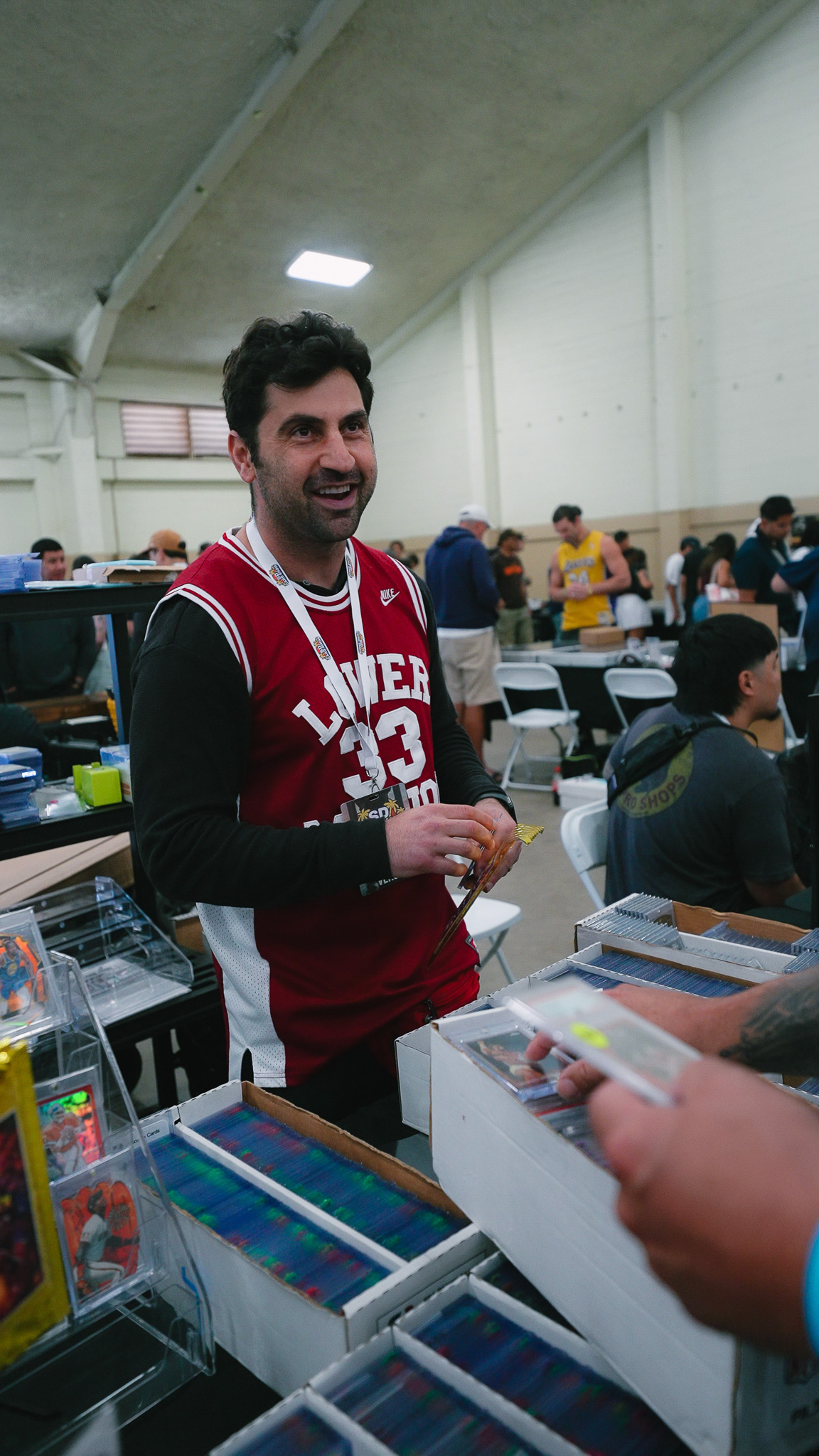 Smiling vendor in a basketball jersey holding a card behind boxes of trading cards